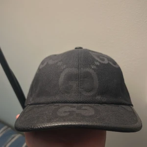 Svart keps från Gucci med GG-mönster - Snygg svart keps från Gucci med diskret GG-mönster över hela kepsen. Justerbart spänne baktill i svart läder och klassisk böjd skärm. Perfekt för dig som gillar stilrena accessoarer med lyxig känsla.