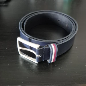 Tommy hilfiger skärp - Snyggt mörkblått bälte från Tommy Hilfiger i äkta skinn. Bältet har ett klassiskt silverfärgat spänne och en detalj med röd, vit och blå rand vid öglan. Perfekt accessoar för att lyfta din outfit. Nypris 500kr