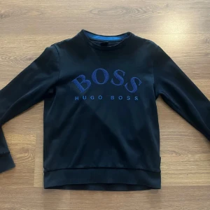 Svart sweatshirt från Hugo Boss - Säljer en svart sweatshirt från Hugo Boss med blå logotyp framtill. Tröjan har rund halsringning och ribbade muddar vid ärmslut och nederkant. Perfekt för en stilren och avslappnad look.