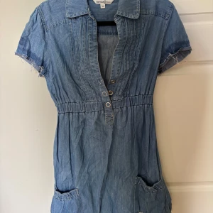 Jeansklänning med kort ärm och knappar - Snygg jeansklänning i blått med korta ärmar och råa kanter. Klänningen har krage, markerad midja och knappar framtill. Två fickor framtill ger en avslappnad look. Har sprättat upp vid armarna själv därav färgskillnaden, super snygg!