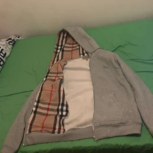 Grå hoodie från Burberry - Grå hoodie från Burberry med dragkedja och ikoniskt beige, svart och röd rutigt foder. Jackan har huva, två fickor fram och ribbade muddar. Tillverkad i bomull med elastan för extra komfort. Använd ett fåtal gånger. Priset är prutat och klart