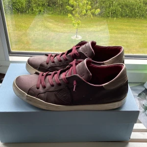 Vinröda sneakers från Philippe Model - Säljer ett par vinröda Philippe Model sneakers med matchande vinröda skosnören och vit sula. Skorna har en klassisk låg modell och är tillverkade i skinn med diskret logga på sidan. Perfekta för dig som gillar stilrena och snygga sneakers. Pris kan sänkas vid snabb affär