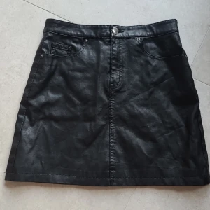 Svart skinnkjol från H&M - Svart kort kjol i skinnimitation. Klassisk femficksmodell med knapp och dragkedja framtill. Snygg och enkel design som passar till många olika stilar.