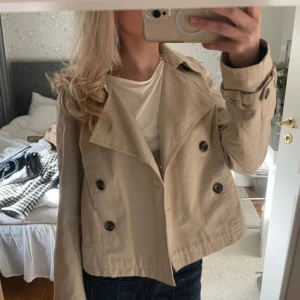 Beige croppad trenchcoat - Säljer en beige croppad trenchcoat med dubbelknäppning och stora slag. Jackan har långa ärmar och snygga detaljer vid ärmsluten. Är köpt från sellpy för 350kr 