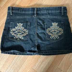  jeanskjol - Mörkblå jeanskjol med snygga broderade detaljer på bakfickorna. Klassisk femficksmodell med silverfärgade knappar och bälteshällor. Perfekta för en avslappnad stil och passar till det mesta.
