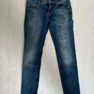 Blå jeans från Jack & Jones - Säljer ett par klassiska blå jeans från Jack & Jones med fem fickor och snygga slitningar. Jeansen har raka ben och normal passform, samt detaljerade sömmar på bakfickorna. Perfekta för en avslappnad stil.