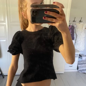 Svart topp med puffärmar och öppen rygg - Trendig svart topp med kort puffärm och ribbad struktur. Toppen har öppen rygg med snygga knytband och rund halsringning. Perfekt för dig som vill ha en stilren men ändå unik look.