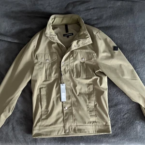 J.Lindeberg jacka - Säljer en stilren grön jacka från J.Lindeberg med klassisk krage och två bröstfickor med lock och knappar. Jackan har dragkedja och tryckknappar framtill samt diskret logotyp på ärmen. Perfekt för kyligare dagar nu i sommar.