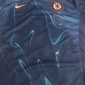 Chelsea FC blå fotbollströja från Nike - Chelsea FC matchtröja i mörkblått med ljusblåa mönster och orange Nike-logga. Klubbloggan sitter på bröstet. Tröjan har en sportig design och är tillverkad i ett lätt och ventilerande material.