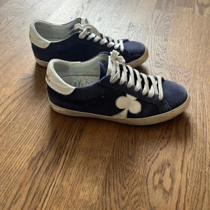 Blå sneakers från The Gallerist  - Säljer ett par blå sneakers från The Dudes med vit klöverdetalj på sidan och vita snören. Skorna har vit hälkappa och platt sula. Perfekta för dig som gillar en unik och stilren look.