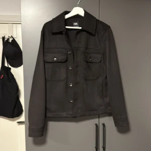 Svart mockajacka från Zara - Säljer en svart mockajacka från Zara med klassisk krage och knäppning framtill. Jackan har två bröstfickor med lock och knappar samt raka ärmar. Snygg och stilren modell som passar till mycket. Passar mig bra och jag är cirka 177
