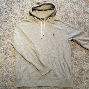 Ralph Lauren hoodie  - Säljer en klassisk grå hoodie från Polo Ralph Lauren med dragsko i huvan och broderad logga på bröstet. Tröjan har känguruficka och är perfekt för en avslappnad stil. Supermjuk och skön att bära.