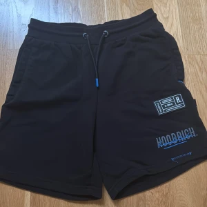 Svarta shorts från Hoodrich med blå detaljer - Svarta shorts från Hoodrich med blå broderade detaljer och tryck på ena benet. Shortsen har snörning i midjan och sidofickor. Tillverkade i mjukt material som passar perfekt för avslappnade dagar.