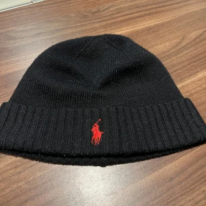 Svart mössa från Polo Ralph Lauren - Svart ribbstickad mössa från Polo Ralph Lauren med röd broderad logotyp framtill. Tillverkad i mjuk merinoull som håller dig varm under kyliga dagar. Klassisk och stilren design. Andvänd 1 gång bara för tyckte inte om att ha mössa på mig. Skick 9.5/10