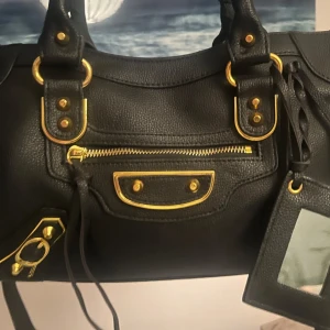 Svart handväska med gulddetaljer - Säljer en svart handväska i skinn med snygga guldfärgade metalldetaljer och dragkedja framtill. Väskan har både handtag och axelrem samt en dekorativ spegel. Perfekt för dig som vill ha en stilren och rymlig accessoar. Köpt på en marknad!