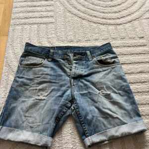 Blå jeansshorts med slitningar - Säljer ett par blå jeansshorts med uppvikta benslut och slitna detaljer framtill. Klassisk femficksmodell med knappgylf. Perfekta för en avslappnad stil under varma dagar.