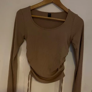 Beige ribbad långärmad topp med knyt - Trendig beige långärmad topp med ribbad struktur och knyt i sidorna för en snygg rynkad effekt. Rund halsringning och figurnära passform. Perfekt att styla till jeans eller kjol.