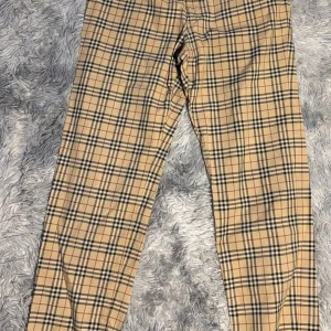 Beige rutiga byxor från Burberry - Snygga beige byxor från Burberry med klassiskt rutigt mönster i svart, vitt och rött. Byxorna har rak passform och är perfekta för dig som gillar en tidlös och trendig stil. Ikoniskt Burberry-mönster som alltid är rätt.