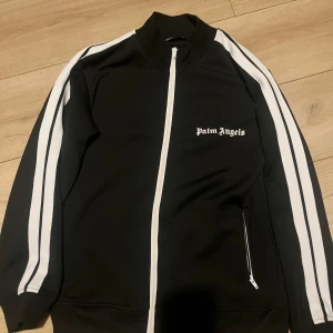 Svart track jacket från Palm Angels - Säljer en svart track jacket från Palm Angels med vita ränder längs ärmarna och dragkedja framtill. Jackan har hög krage och logga på bröstet. Perfekt för en sportig och trendig look.