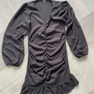Svart ribbad klänning med volang - Svart klänning med ribbad struktur, långa puffärmar och v-ringning. Klänningen har rynkade detaljer framtill och en volangkant nertill för en trendig look. Perfekt för dig som gillar stilrena och moderna plagg.