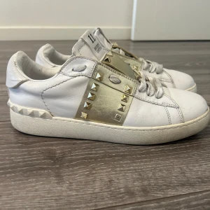  Valentino med gulddetaljer och nitar - Säljer ett par vita Valentino sneakers med guldfärgad rem och ikoniska nitar i guld längs sidan och bak på sulan. Skorna har snörning och en platt sula. Perfekta för dig som gillar lyxiga detaljer och streetstyle. (Storlek:42)
