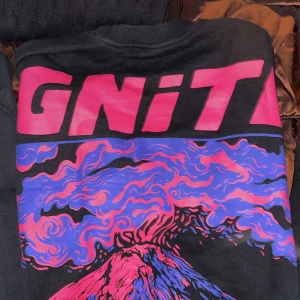 Svart sweatshirts med färgglatt vulkantryck - Cool svart t-shirt med stort tryck av en vulkan i rosa, blått och lila. Texten 'IGNITE' i rosa upptill. Perfekt statement-plagg för dig som gillar unika motiv och färg. Rund halsringning och normal passform.