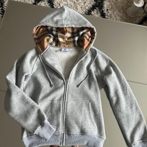 Grå hoodie med Burberry-rutig huva - Grå hoodie från Burberry med dragkedja och klassisk Burberry-rutig insida på huvan. Mjuk bomullskänsla och ribbade muddar. Perfekt för en stilren och avslappnad look. Står M men sitter som S
