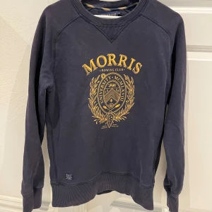Mörkblå sweatshirt från Morris R. Club - Säljer en mörkblå sweatshirt från Morris R. Club med guldfärgat tryck och emblem på bröstet. Tröjan har långa ärmar, rund halsringning och ribbade muddar vid ärmslut och nederkant.