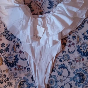 Vit volangbody från Zara - Supersnygg vit body från Zara med breda volangdetaljer över axlarna och ribbad struktur. Perfekt för dig som vill sticka ut på stranden med en trendig och feminin look. Hög benskärning och djup urringning framtill. PRIS KAN DISKUTERAS.