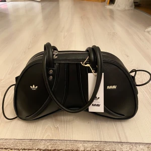 Svart handväska från Adidas x AVAVAV - Unik svart handväska i samarbete mellan Adidas och AVAVAV Nypris 6800kr. Väskan har silvriga dragkedjor, två handtag och axelrem. Tillverkad i syntetmaterial med Adidas-logga och AVAVAV-detaljer på framsidan. Perfekt för dig som vill sticka ut med en cool accessoar.