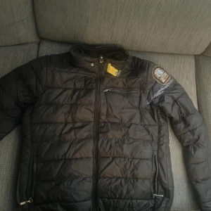 Svart dunjacka från Parajumpers - Säljer en svart dunjacka från Parajumpers med patch på ärmen och dragkedja framtill. Jackan är superlätt och har ståkrage samt fickor med dragkedja. Perfekt för dig som vill ha en lätt och smidig jacka med snygg design. Priser är ej hugget i sten.