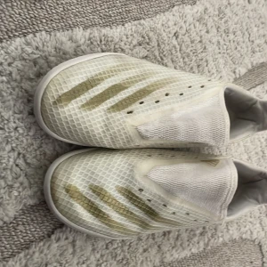 Vita fotbollsskor från Adidas - Säljer ett par vita junior fotbollsskor från Adidas med guldiga detaljer och klassiska tre ränder på sidan. Skorna har en slät ovandel utan snörning och är designade för optimal bollkontroll. Perfekta för dig som vill sticka ut på planen.