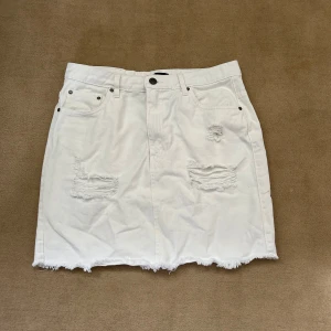 Vit jeanskjol med slitna detaljer - Vit jeanskjol från NELLY med slitna detaljer framtill och rå kant nertill. Kjolen har klassiska fem fickor och knappstängning. Perfekt för en avslappnad och trendig look.