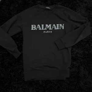 Svart sweatshirt från Balmain Paris - Säljer en svart sweatshirt från Balmain Paris med vit logotyp på bröstet. Tröjan har rund halsringning och långa ärmar. Perfekt för en stilren och trendig look.