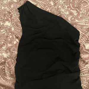 Svart one shoulder klänning - Säljer en svart topp med one shoulder-design. toppen har en stilren och enkel form som passar perfekt för dig som gillar minimalistisk stil. Materialet känns mjukt och följsamt mot kroppen.