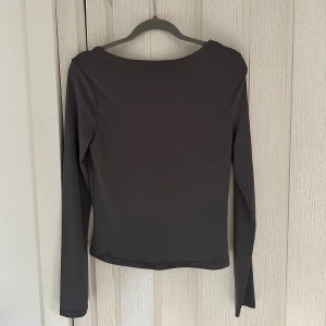 Mörkgrå långärmad topp med djup urringning i ryggen - Säljer en stilren mörkgrå långärmad topp med djup urringning i ryggen. Toppen har en enkel och slät design, perfekt för att matcha med olika outfits. Materialet känns mjukt och stretchigt för en bekväm passform.