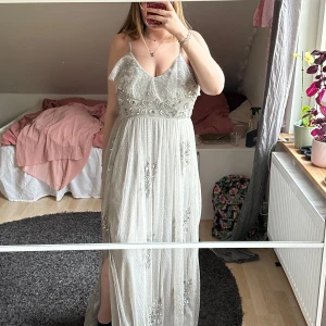 Ljusgrå långklänning med paljetter från Maya Deluxe - Säljer en ljusgrå långklänning från Maya Deluxe med smala axelband och v-ringning. Klänningen är dekorerad med glittriga paljetter och har en lätt transparent mesh med broderade detaljer. Perfekt för speciella tillfällen.