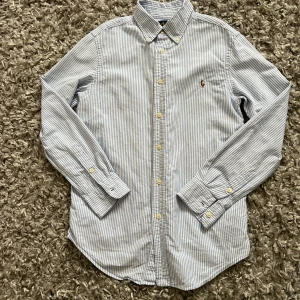 Blårandig skjorta från Ralph Lauren - Klassisk blårandig skjorta från Ralph Lauren med broderad logga på bröstet. Skjortan har lång ärm, knappar framtill och button-down krage. Perfekt för en stilren och tidlös look.