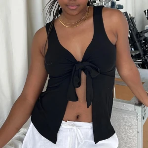 Svart knytlinne  - Säljer ett svart ärmlöst linne med djup v-ringning och knyt framtill. Toppen har en croppad passform och är perfekt för varma dagar. Enkel och stilren design som passar till mycket.