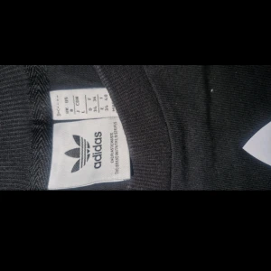 Svart klänning från Adidas Originals - Svart t-shirt från Adidas Originals med stort vitt logotryck på bröstet och klassiska vita ränder på axeln. Tillverkad i mjuk bomull och har en normal passform med korta ärmar. Perfekt för en sportig och avslappnad stil.