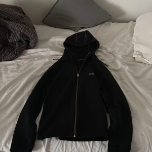 Svart hoodie med dragkedja från BOSS - Säljer en svart hoodie från BOSS med dragkedja framtill och huva med snörning. Tröjan har diskreta vita sömmar på huvan och BOSS-logga broderad på bröstet. Perfekt för en avslappnad och stilren look.