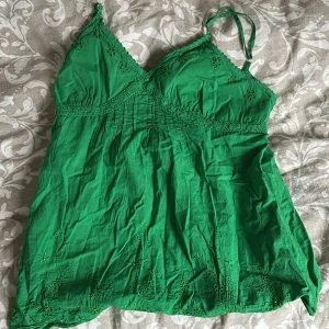 Grön linnetopp med broderade blommor - Säljer en grön linnetopp med tunna axelband och fina broderade blommor framtill och nertill. Toppen har en lätt v-ringning och smock under bysten för en bekväm passform. Perfekt för varma dagar! Passar någon som gillar en lämgre passform, säljer pga för stor💓