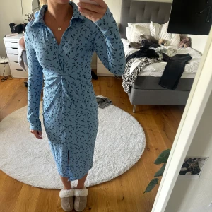 Blå blommig långärmad klänning med krage - Säljer en ljusblå långärmad klänning med svart blommigt mönster och krage. Klänningen har en v-ringning, åtsittande passform och en liten slits framtill. Klänningen är endast använd ett fåtal gånger. Den är från Gina tricot och är i storlek S