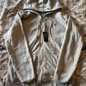 Grå hoodie från Polo Ralph Lauren - Säljer en klassisk grå hoodie från Polo Ralph Lauren med dragkedja och huva. Tröjan har långa ärmar, fickor framtill och den ikoniska loggan broderad på bröstet. Perfekt för en avslappnad stil.