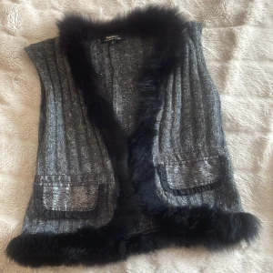 Grå stickad väst med fuskpälsdetaljer - Säljer en grå stickad väst med svarta fuskpälsdetaljer längs krage och nederkant. Västen har två fickor framtill, dekorativa knappar bak och stängs med hyskor. Perfekt för att lyfta en outfit!
