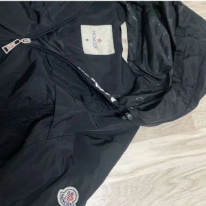 Svart vindjacka från Moncler med huva - Säljer en svart vindjacka från Moncler med huva och dragkedja framtill. Jackan har Moncler-logga på ärmen och tryckt Moncler-text längs insidan av huvan. Perfekt för dig som vill ha en stilren och sportig jacka.