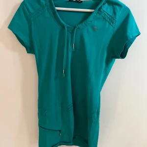 Turkos lång t-shirt hoodie - Turkos tshirt med sportig känsla från Extory. Kortärmad modell med huva, dragsko i halsen och dekorativa sömmar samt mönsterdetaljer. Praktisk ficka med dragkedja framtill. Perfekt för en avslappnad och aktiv stil. Står XL på lappen men skulle säga att den sitter mer som en S💞