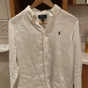 Vit linneskjorta från Ralph Lauren - Säljer en stilren vit linneskjorta från Ralph Lauren med klassisk krage och knappar framtill. Skjortan har en broderad logga på bröstet och långa ärmar. Perfekt för en avslappnad men ändå elegant look. Priset är inte hugget i sten