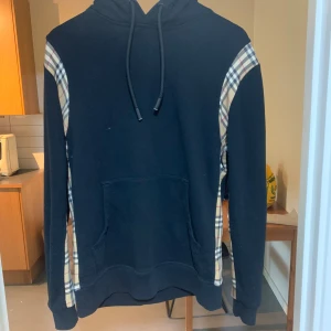 Svart hoodie från Burberry med rutiga detaljer - Säljer en svart hoodie från Burberry med klassiska beige, röda och svarta rutiga paneler längs ärmarna. Tröjan har huva med dragsko och en stor magficka framtill. Tillverkad i mjuk bomull för en skön känsla.