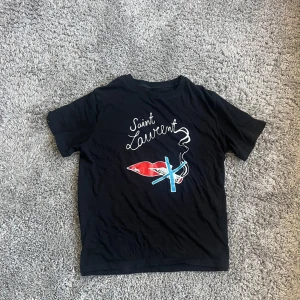 Svart t-shirt med tryck från Saint Laurent - Snygg svart t-shirt från Saint Laurent med ett coolt tryck av en röd läpp och blå cigrett. Tröjan är i bra skick men kan ej garantera äkta het, passar s-m